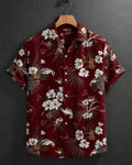A short-sleeve button-up camp shirt with a red-burgundy base and a white, beige, and brown tropical floral print, hanging on a wooden hanger against a dark gray textured wall.