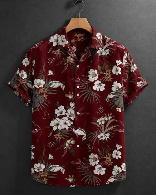 A short-sleeve button-up camp shirt with a red-burgundy base and a white, beige, and brown tropical floral print, hanging on a wooden hanger against a dark gray textured wall.