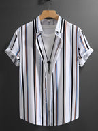 A men's short-sleeve button-up shirt with vertical stripes in black, white, blue, and brown, styled open over a white t-shirt on a wooden hanger against a dark gray wall.