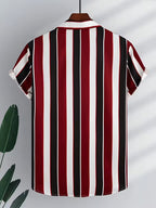 A short-sleeve collared shirt hanging on a wooden hanger, featuring bold vertical stripes in red, black, and white. The shirt is displayed against a light gray background with soft leaf shadows and a decorative green plant leaf on the left.