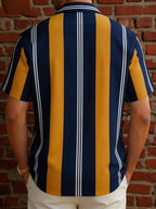 A man viewed from the back wearing a short-sleeve button-up shirt with bold vertical stripes in navy blue, mustard yellow, and thin white stripes, standing against a red brick wall.