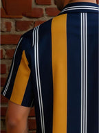 A close-up back view of a person wearing a short-sleeve collared shirt with bold vertical stripes in navy blue, mustard yellow, and white, set against a red brick wall background.
