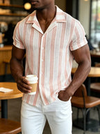 A man wearing a short-sleeve button-up shirt with vertical pink and white stripes, holding a paper coffee cup in a cafe setting.