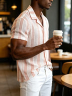 A man in a cafe wearing a short-sleeve button-up shirt with vertical pink and white textured stripes, paired with white pants, holding a paper coffee cup.
