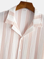 A close-up of a textured knit button-up shirt with vertical stripes in pale pink and white, hanging on a wooden hanger against a light gray background.