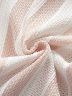 A close-up of a soft knit fabric with a textured openwork pattern in pale blush pink and white, arranged in gentle folds.