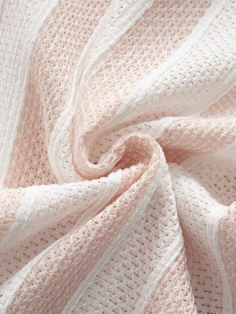 A close-up of a soft knit fabric with a textured openwork pattern in pale blush pink and white, arranged in gentle folds.