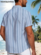 A man standing on a beach wearing a light blue and white vertically striped short-sleeve button-down shirt, shown from the back. Palm trees and ocean are visible in the background.