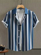 A men's short-sleeve button-up shirt with vertical blue, navy, and white stripes, styled open over a white t-shirt on a wooden hanger against a textured gray wall.