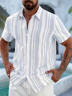 A man wearing a short-sleeve button-up shirt with vertical white, light blue, and gray stripes, paired with white pants, standing outdoors near a pool.