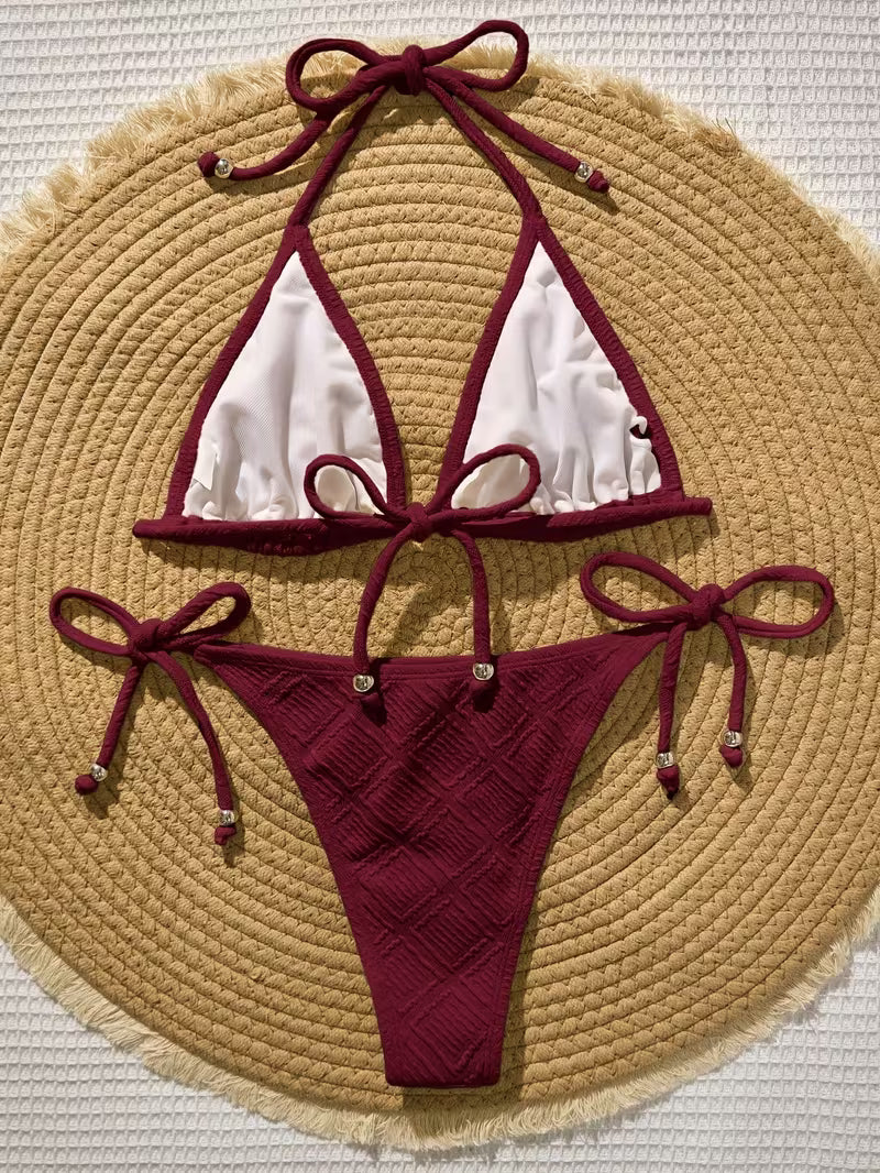A two-piece string bikini laid flat on a round woven straw hat. The bikini has deep burgundy ties and trim with white mesh-style front panels, and small metallic bead accents at the tie ends.