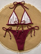 A two-piece string bikini laid flat on a round woven straw hat. The bikini has deep burgundy ties and trim with white mesh-style front panels, and small metallic bead accents at the tie ends.
