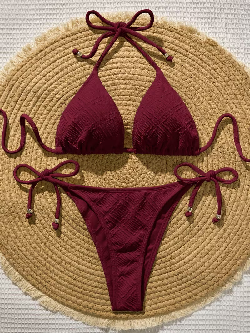 A two-piece bikini set in deep burgundy with a textured, wavy knit pattern, featuring halter top straps and side-tie bottoms, laid flat on a round woven straw mat.
