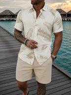 A man wearing a cream short-sleeve button-up shirt with a beige palm leaf pattern and matching beige shorts, standing on a wooden boardwalk next to turquoise water with overwater bungalows in the background.