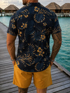 A man viewed from behind wearing a navy blue short-sleeve button-up shirt with a yellow tropical floral and palm leaf print, paired with mustard yellow shorts, standing on a wooden boardwalk next to overwater bungalows and turquoise water.