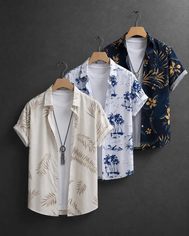 Three men's short-sleeve button-up shirts with tropical prints are displayed on wooden hangers against a dark gray wall. The shirt on the left is cream with beige palm leaf prints, the middle is white with blue palm tree prints, and the right is navy blue with yellow tropical floral prints.