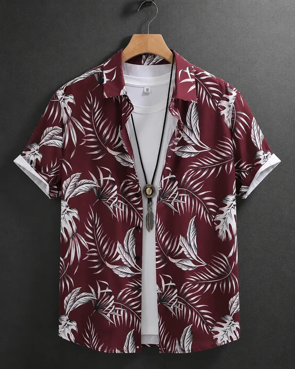 A short-sleeve button-up shirt with a white tropical palm leaf pattern on a deep burgundy background, hanging on a wooden hanger over a white t-shirt against a dark gray background.