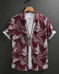A short-sleeve button-up shirt with a white tropical palm leaf pattern on a deep burgundy background, hanging on a wooden hanger over a white t-shirt against a dark gray background.