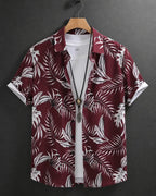 A short-sleeve button-up shirt with a white tropical palm leaf pattern on a deep burgundy background, hanging on a wooden hanger over a white t-shirt against a dark gray background.