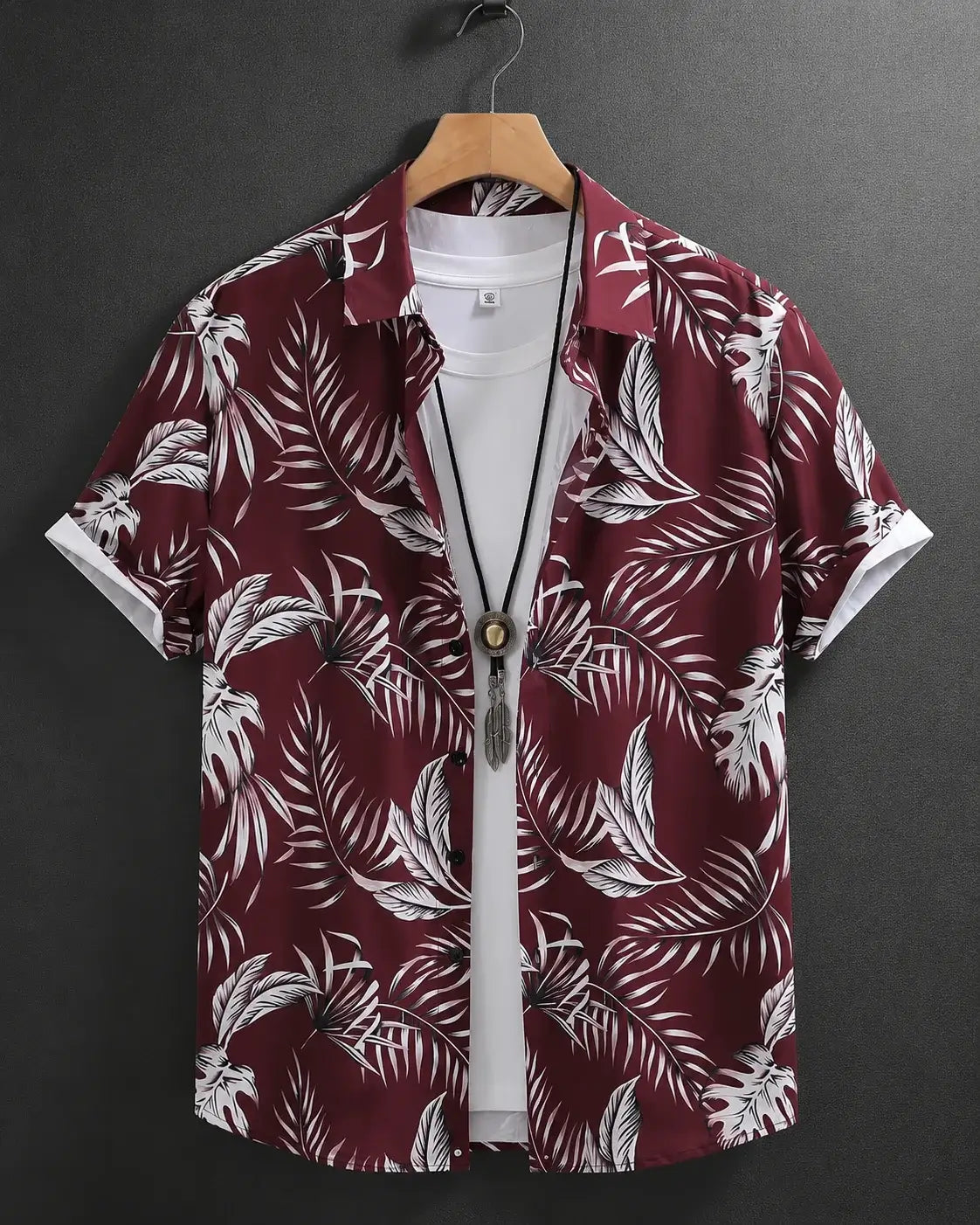 A short-sleeve button-up shirt with a white tropical palm leaf pattern on a deep burgundy background, hanging on a wooden hanger over a white t-shirt against a dark gray background.