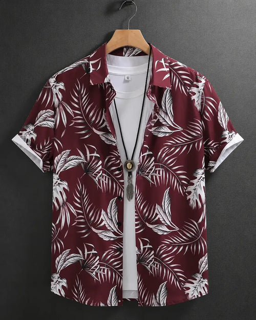 A short-sleeve button-up shirt with a white tropical palm leaf pattern on a deep burgundy background, hanging on a wooden hanger over a white t-shirt against a dark gray background.