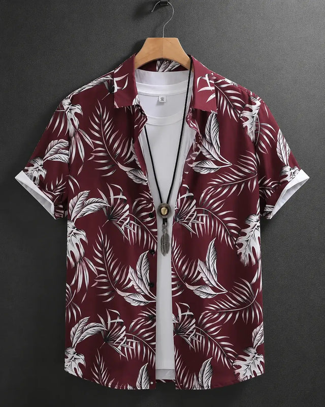 A short-sleeve button-up shirt with a white tropical palm leaf pattern on a deep burgundy background, hanging on a wooden hanger over a white t-shirt against a dark gray background.