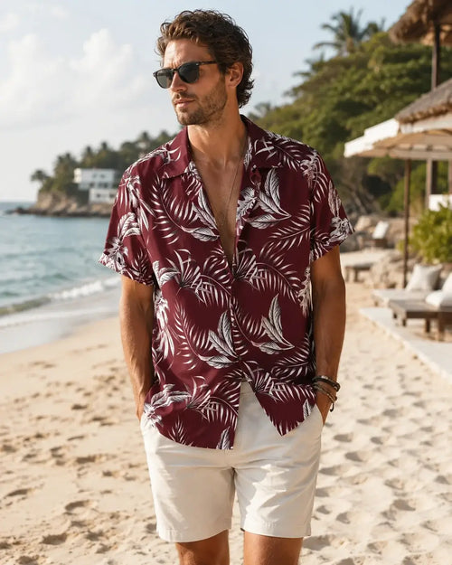 A man wearing a burgundy short-sleeve button-up shirt with a white palm leaf pattern, styled with off-white shorts on a sandy beach.