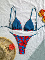 A two-piece bikini set laid flat on a white fabric background, styled with a straw hat, orange flower, and yellow-orange swim goggles. The bikini top is blue with thin red and white stripes and red side accents, while the matching bottoms feature a bold red, blue, and orange floral print.