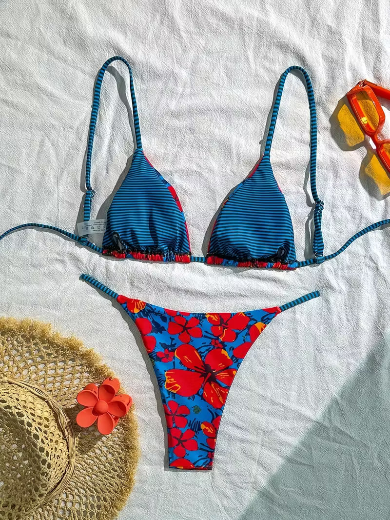 A two-piece bikini set laid flat on a white fabric background, styled with a straw hat, orange flower, and yellow-orange swim goggles. The bikini top is blue with thin red and white stripes and red side accents, while the matching bottoms feature a bold red, blue, and orange floral print.