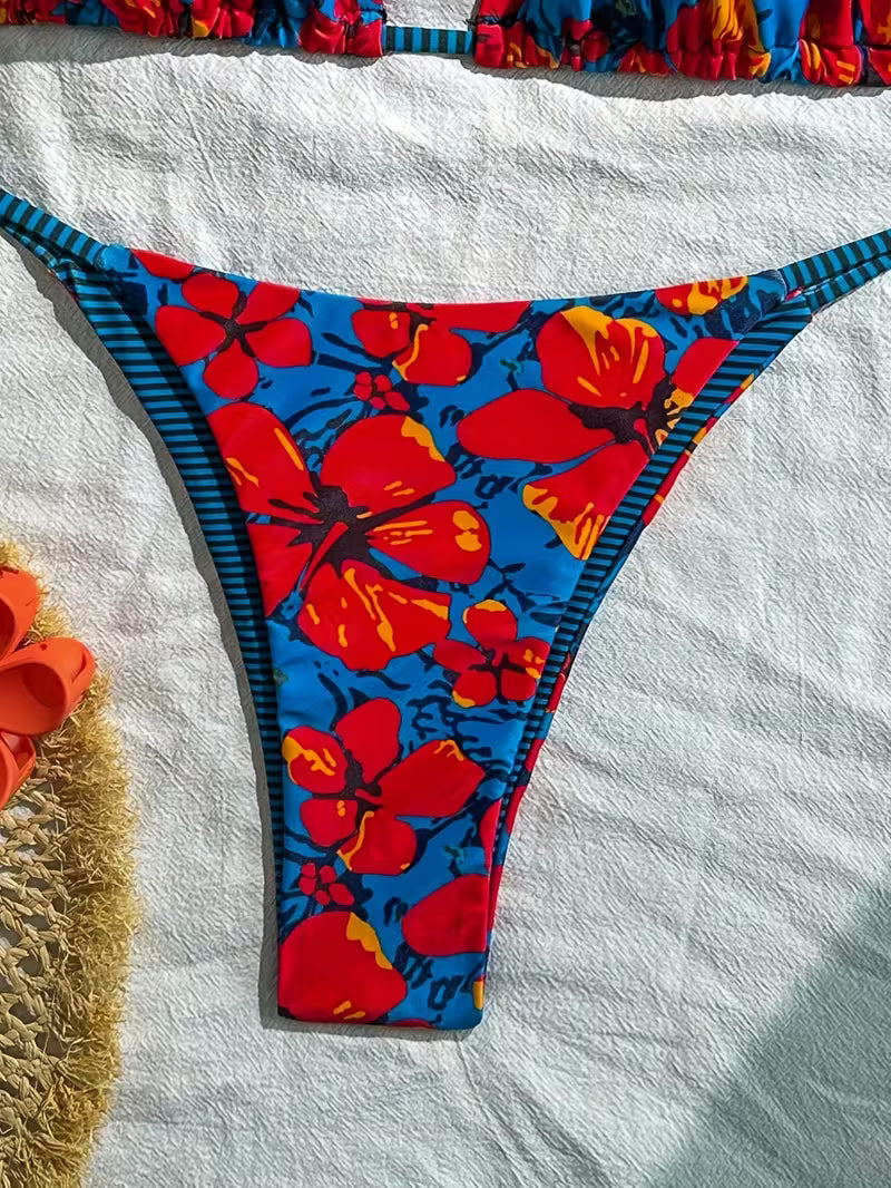 A pair of women's bikini bottoms laid flat on a white textured towel. The bottoms feature a vibrant blue base with a bold red and orange hibiscus floral print, accented by dark blue and orange striped side straps.