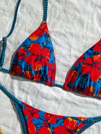 A close-up flat lay of a bikini top and bottom with a vibrant blue base, large red hibiscus floral print, and dark blue and black striped straps, laid on a textured white towel.
