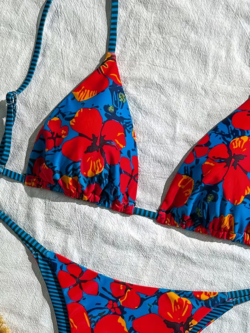 A close-up flat lay of a bikini top and bottom with a vibrant blue base, large red hibiscus floral print, and dark blue and black striped straps, laid on a textured white towel.