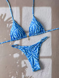 A blue and white patterned string bikini set laid flat on a textured beige wall with soft window shadow overlays. The set includes a triangle bikini top with thin side straps and a matching low-rise bikini bottom.