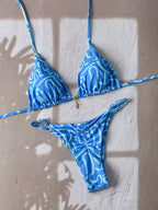 A blue and white patterned string bikini set laid flat on a textured beige wall with soft window shadow overlays. The set includes a triangle bikini top with thin side straps and a matching low-rise bikini bottom.