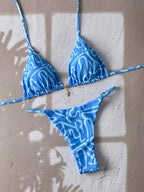 Blue and white patterned string bikini set laid flat on a textured beige wall with soft window shadow overlays.