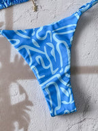 Blue thong-style bikini bottom with a white abstract wavy pattern, laid flat on a textured light gray surface.