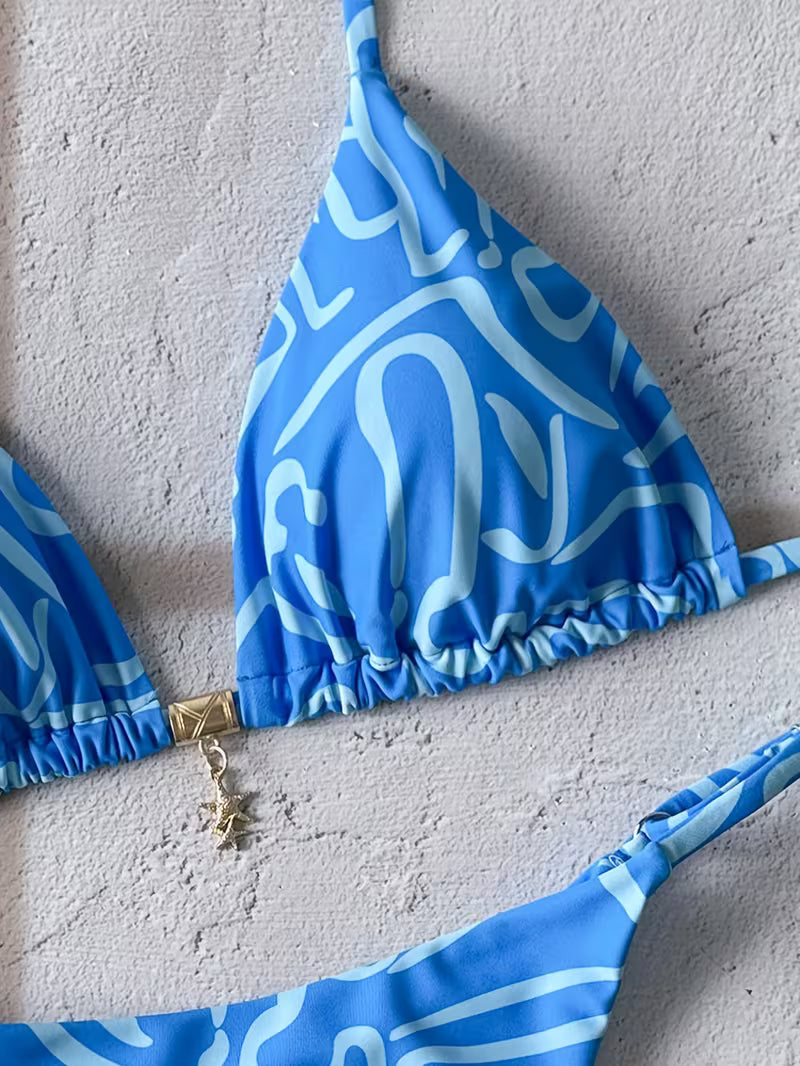 A close-up of a blue bikini top with a white abstract wavy pattern and a small gold charm, laid flat on a textured light gray surface.