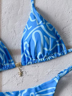A close-up of a blue bikini top with a white abstract wavy pattern and a small gold charm, laid flat on a textured light gray surface.