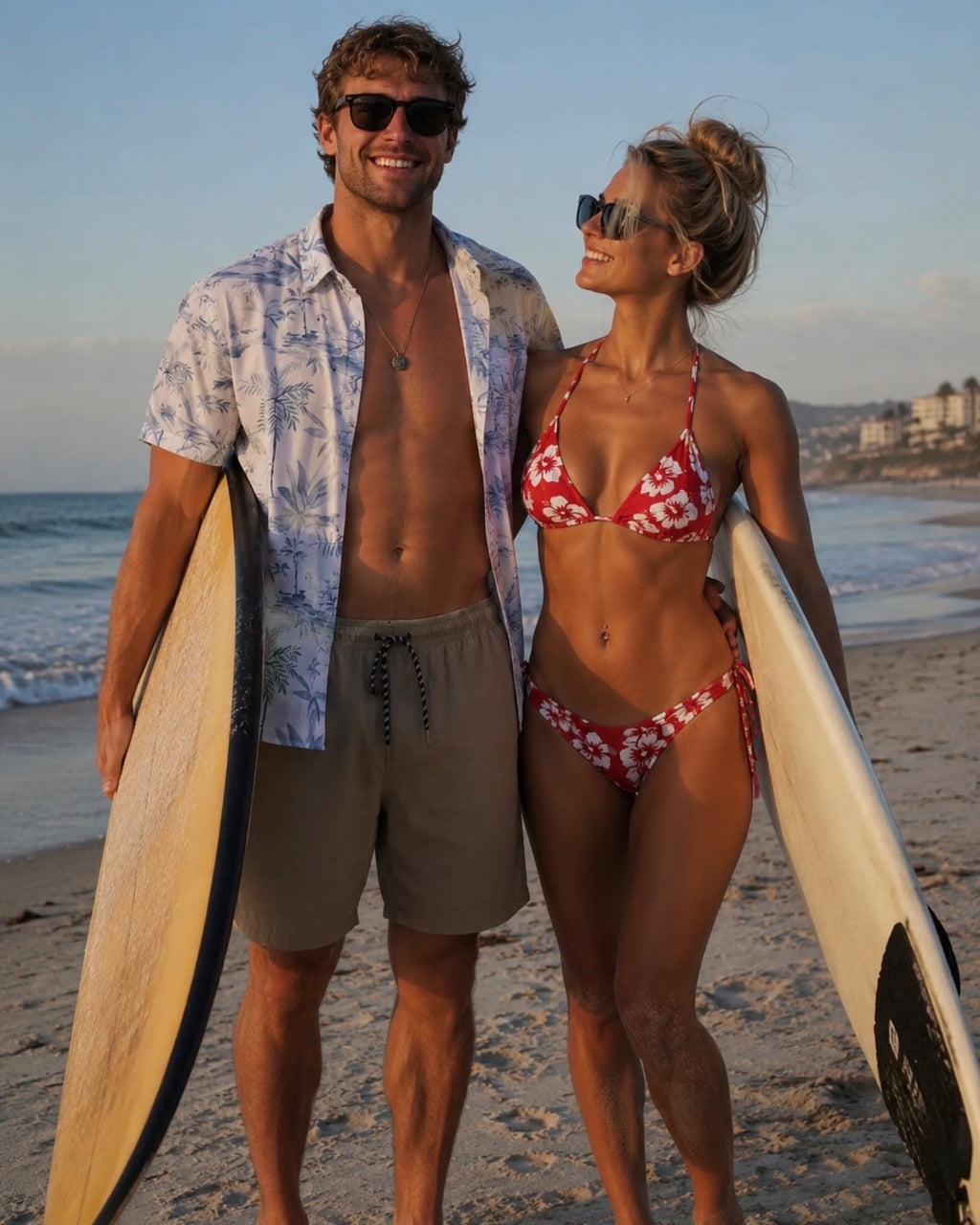 A couple on a sandy beach at sunset, each holding a surfboard. The man wears a light blue patterned short-sleeve button-up shirt, beige shorts, and sunglasses.