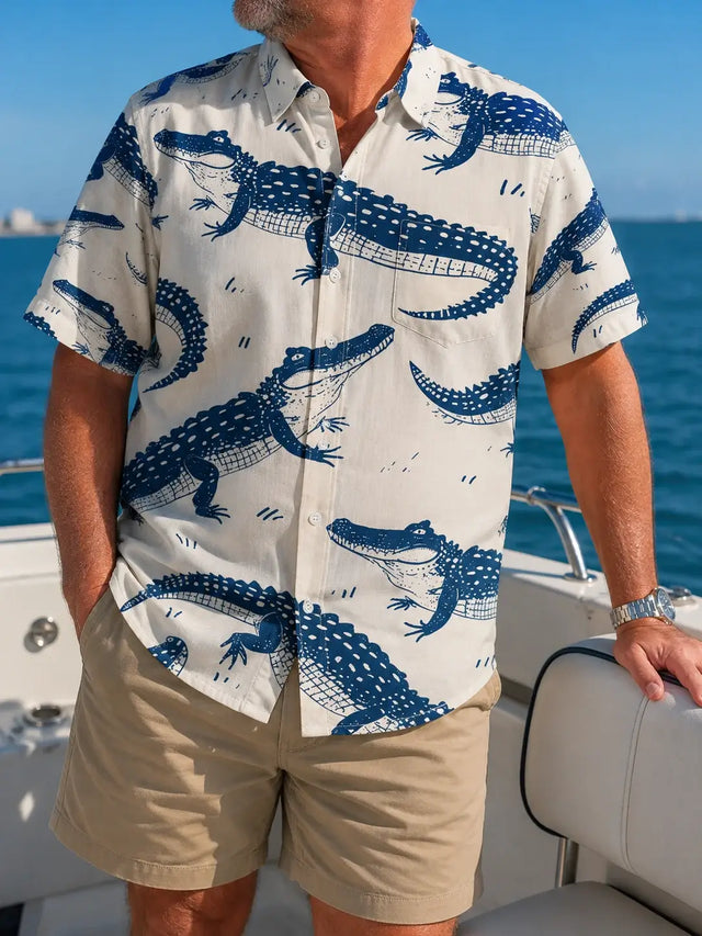 A man wearing a short-sleeve button-up shirt with a blue alligator print on a cream background, paired with khaki shorts, standing on a boat with the ocean in the background.