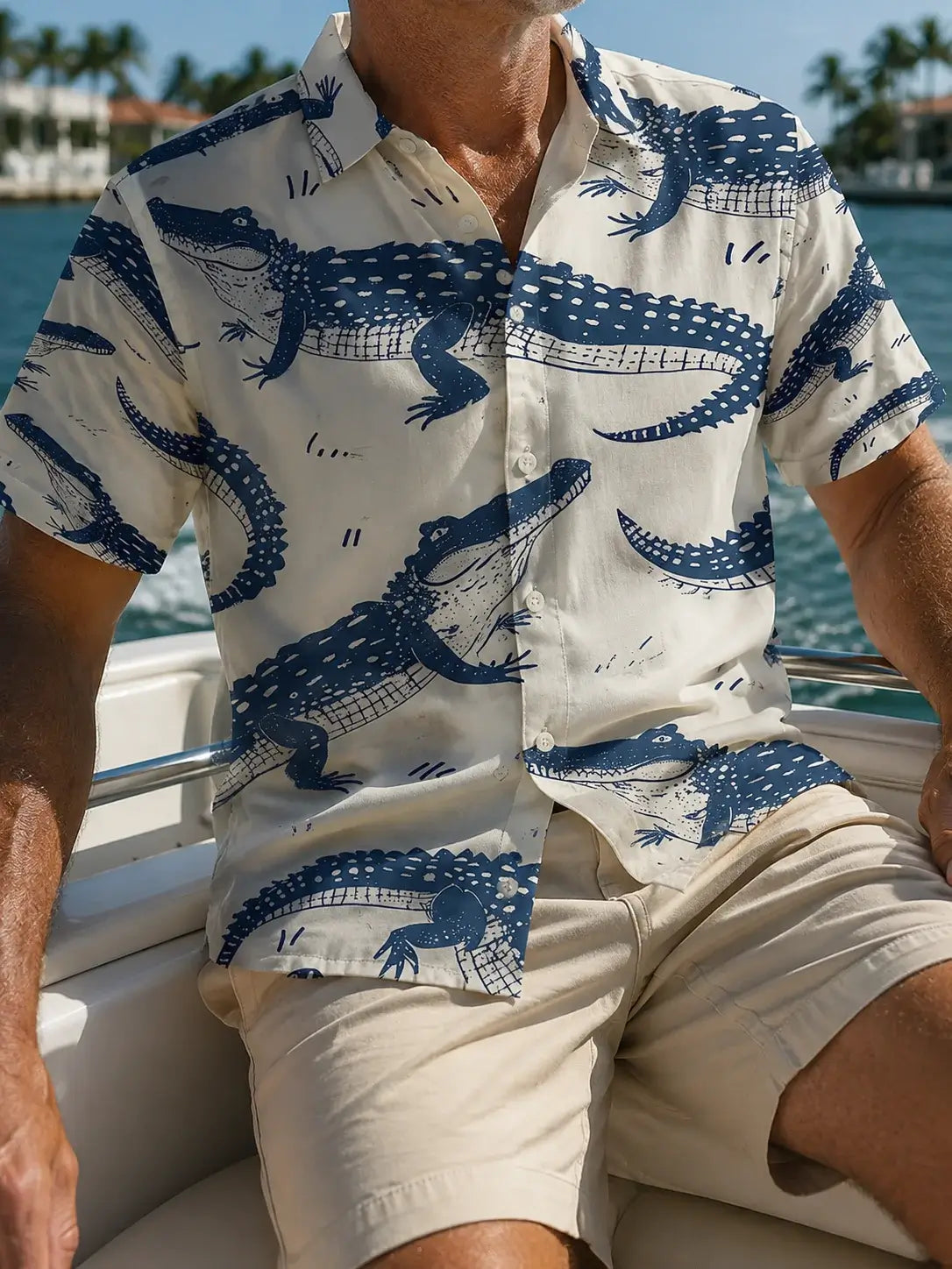 A man wearing a short-sleeve button-up shirt with a blue alligator print on an off-white background, paired with beige shorts while sitting on a boat.