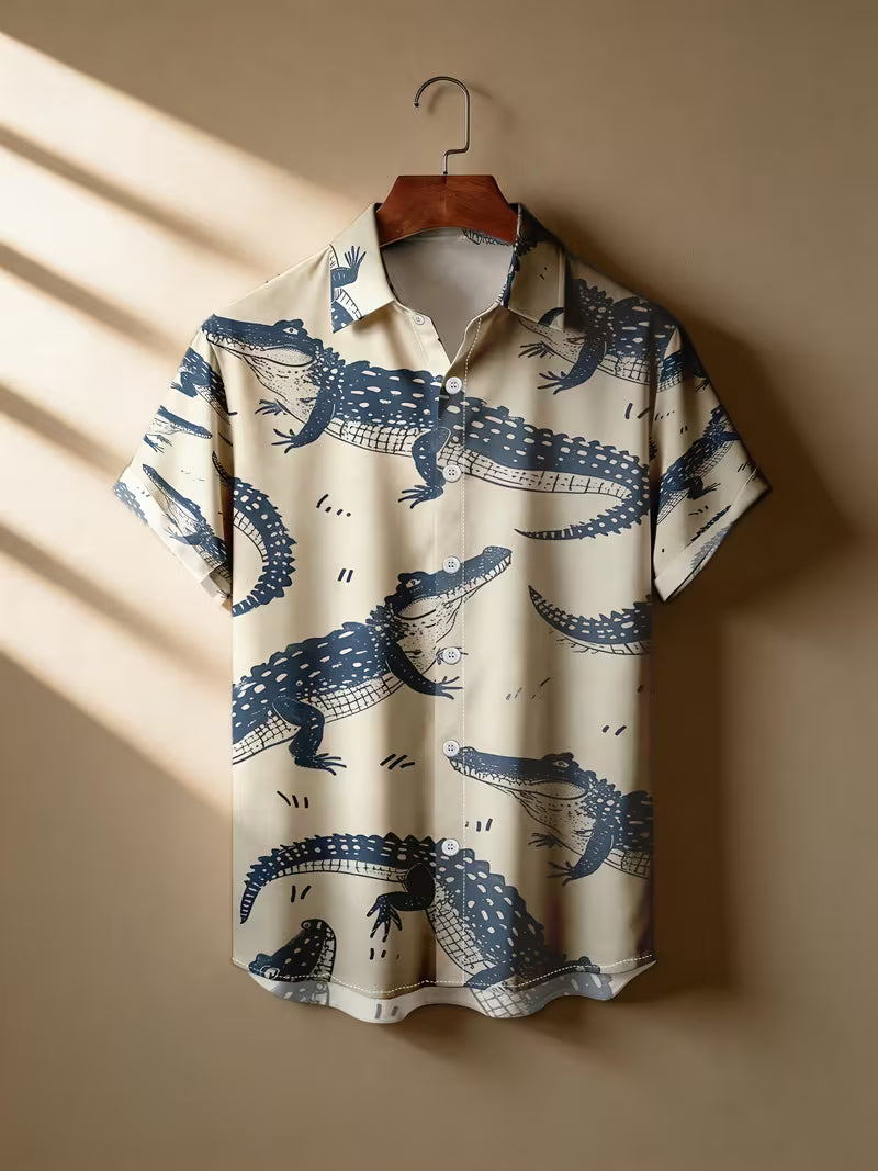 A short-sleeve camp collar button-up shirt with an all-over blue alligator print on a cream background, hanging on a wooden hanger against a warm beige wall with diagonal sunlight shadows.