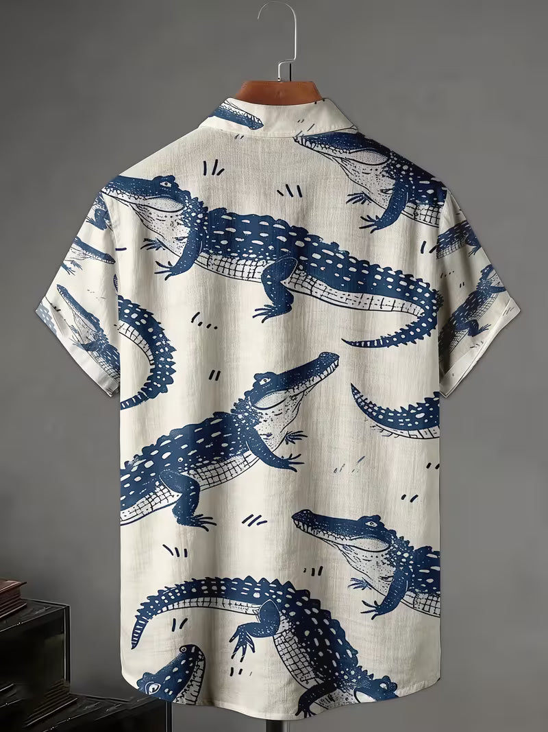 A short-sleeve camp collar shirt with an all-over blue alligator print on a cream-colored linen-like fabric, displayed on a wooden hanger against a dark gray background.