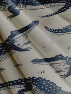 A close-up of a fabric or wallpaper pattern featuring dark navy blue alligators printed on a warm cream background.