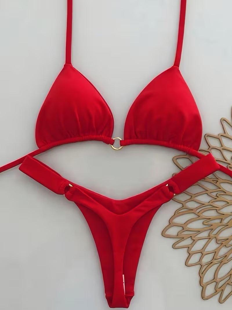 A bright red string bikini set with a triangle top and matching low-rise bottoms, featuring small gold metal ring accents at the center of the top and on the side ties, displayed against a light background with a decorative woven mat on the right.