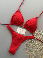 A red two-piece bikini set with a triangle top and matching thong bottom, laid flat on a gray background with a small handwritten note and a decorative gold fan.