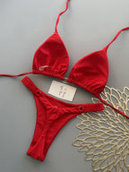 A red string bikini set laid flat on a gray background, including a triangle top with side ties and matching low-rise bottoms. A small handwritten price tag is placed on the bikini, and a decorative gold fan is partially visible on the right.