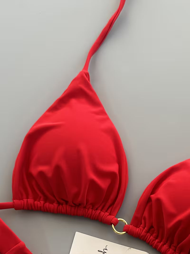 A close-up of a bright red triangle bikini top with a thin shoulder strap, gathered elastic band, and a small gold ring detail, laid flat on a light gray background.