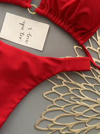 A red bikini top and bottom laid flat on a gray surface, styled with a gold scalloped decorative prop and a small handwritten note card.