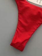 A close-up of a bright red garment, likely a bikini bottom or high-cut swim brief, laid flat on a neutral gray background with a white paper tag partially visible at the top.